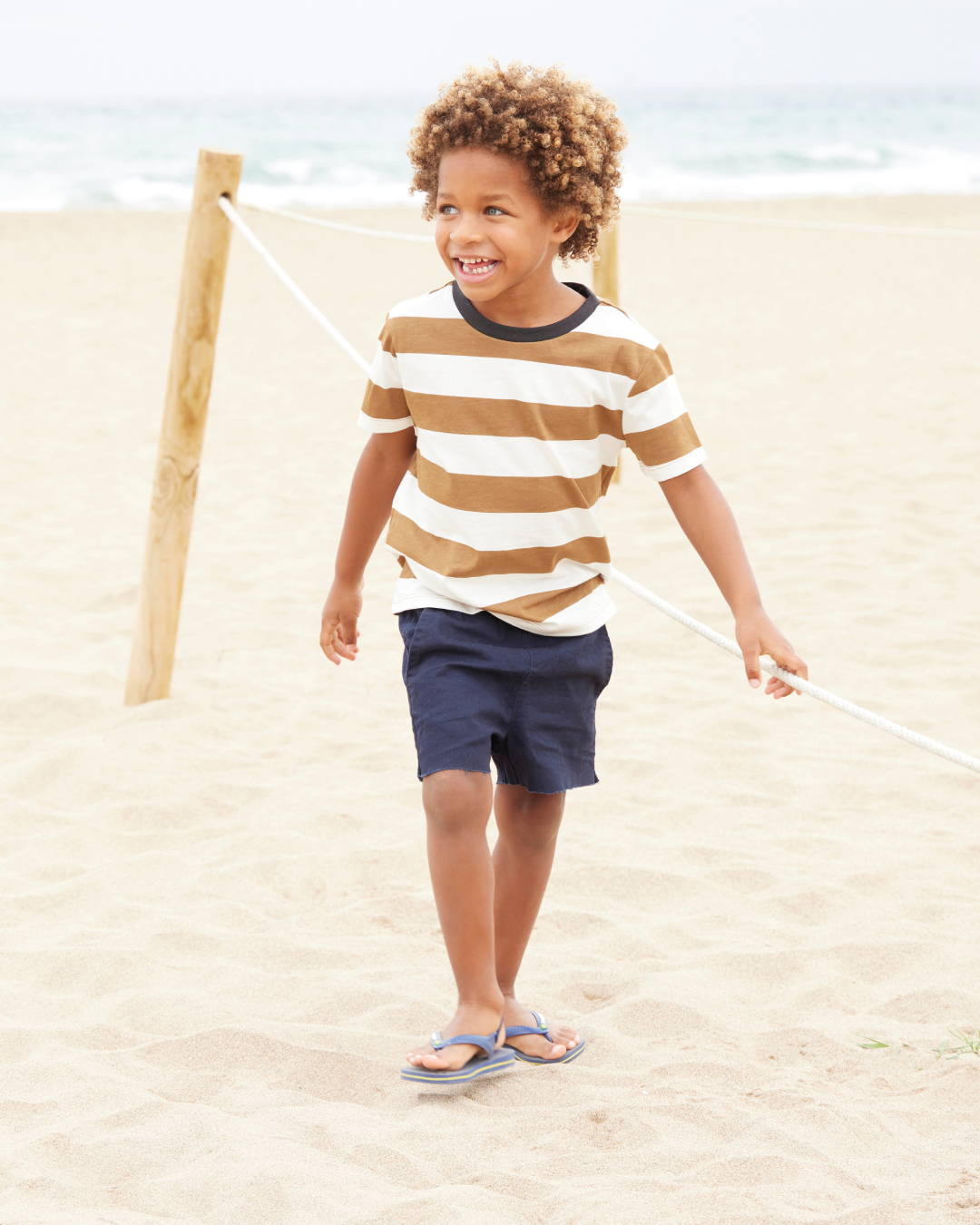 little boy walking on beach, wearing a beige and white striped shirt, navy shorts, and blue havaianas flip flops