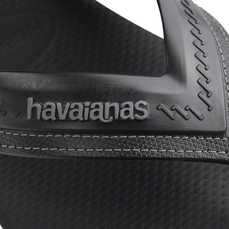 black sandals with rice patterned grooves and striped grooves on the footbed, with black straps and a grey stitched like pattern, zoomed into light grey havaianas logo on strap