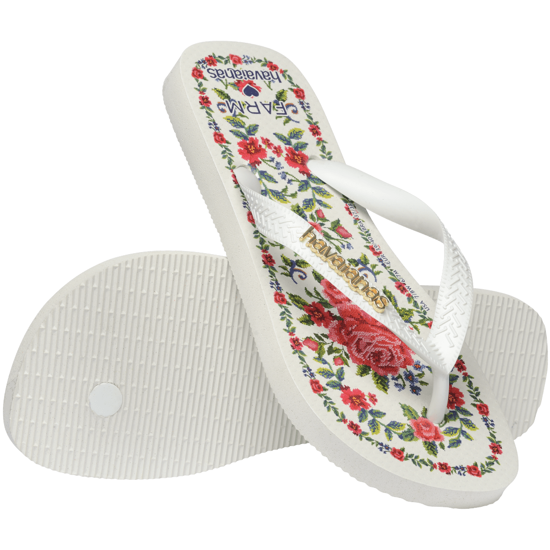 flip flops with pink flowers on a white background with white straps with a gold havaianas logo on them, one flip flop resting on the other, toe side facing down