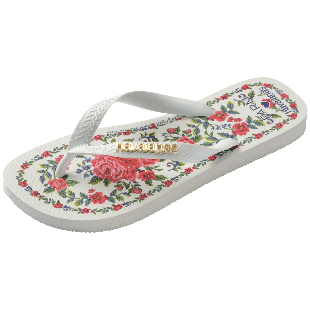 flip flops with pink flowers on a white background with white straps with a gold havaianas logo on them, front-diagonal facing